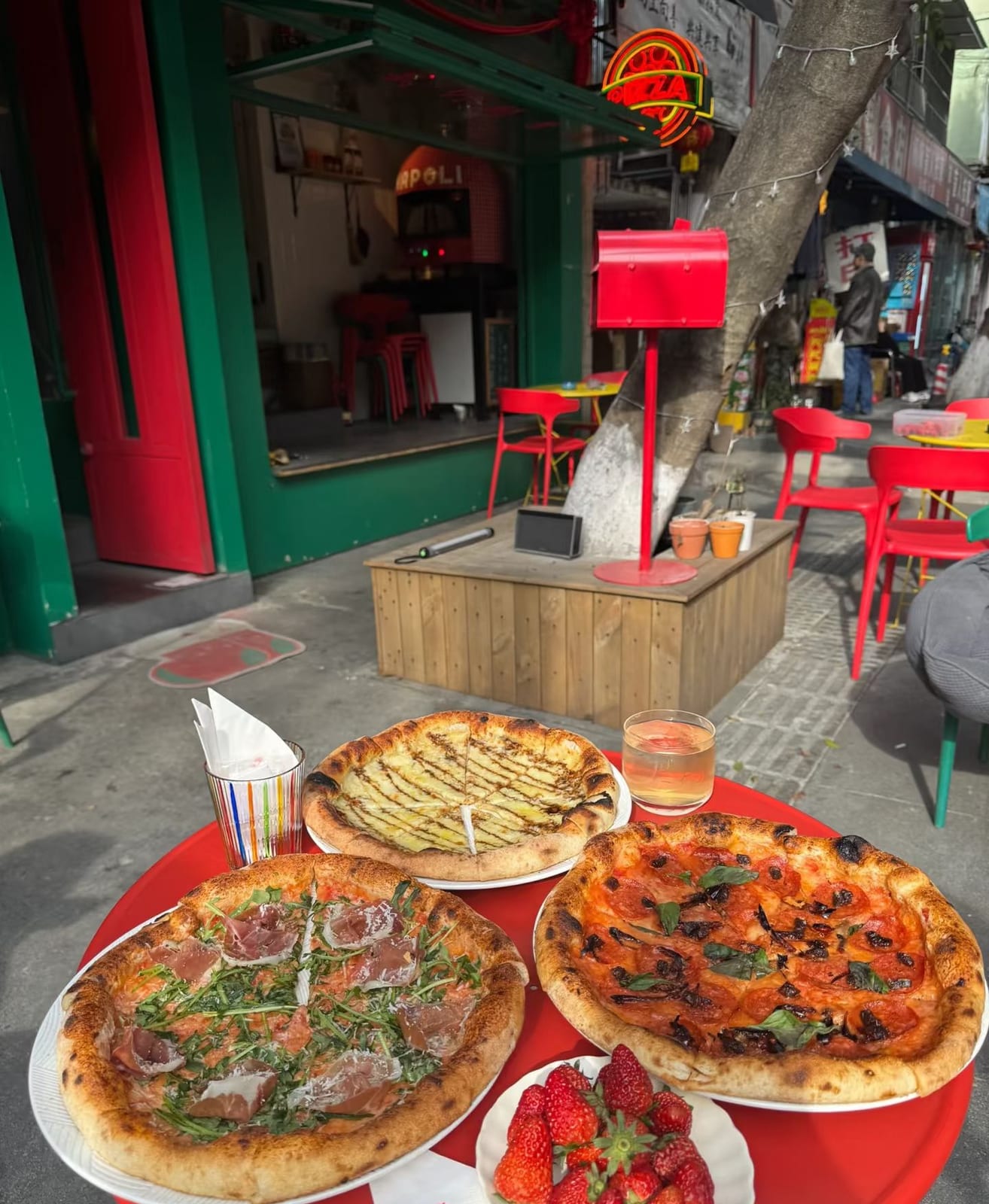 窄门红绿撞色门头全景:NAPOLI 窑炉在店内,街边红桌上摆着三张披萨和一盘草莓